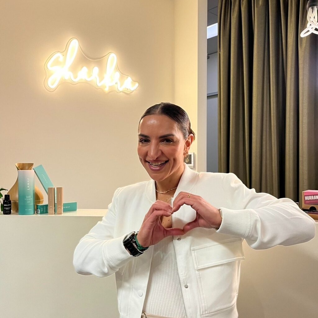 Tanya is dressed all in white and has made a heart shape with her hands. She smiles at the camera and stands in front of her reception desk with a Sharkra neon sign lighting in the background